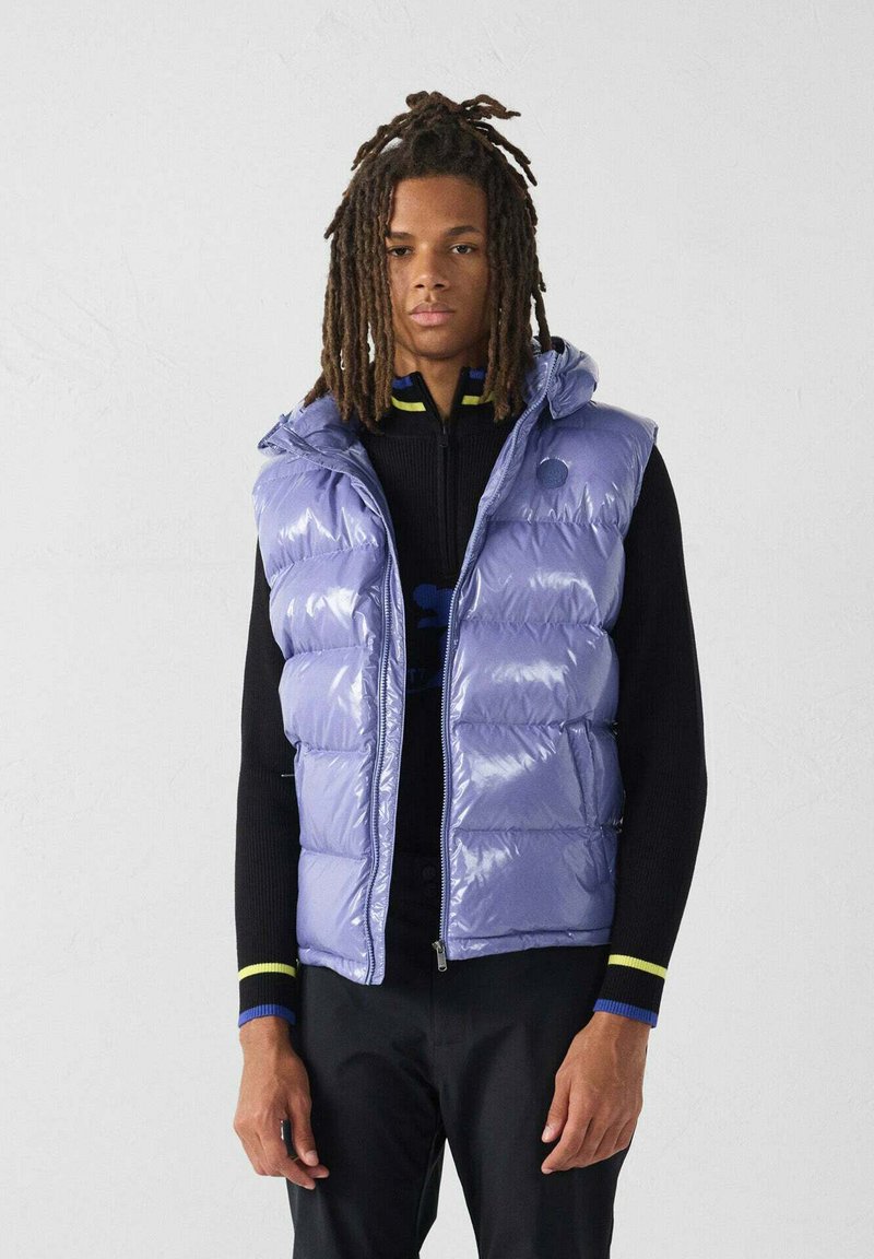 Lichtpaarse puffer-vest met een glanzende afwerking, voorzien van een capuchon, ritssluiting en meerdere horizontale doorgestikte secties. Gedragen over een zwart shirt.