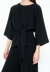 Top nero realizzato in un tessuto liscio. Presenta un collo rotondo, maniche ampie e un dettaglio di nodo frontale in vita per un aspetto aderente.