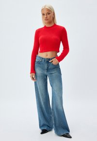 Top rosso, peloso, a maniche lunghe accorciate abbinato a jeans azzurri chiari a vita alta e scarpe nere con punta a spillo.