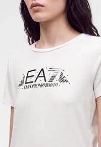 Γυναίκα που φοράει άσπρο μπλουζάκι με μαύρο λογότυπο EA7 Emporio Armani στο στήθος, που δείχνει τον λαιμό, τους ώμους και το πάνω μέρος του κορμού.