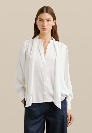 Mujer joven con blusa blanca de manga larga con detalle de bufanda y jeans de color azul oscuro, de pie contra un fondo beige liso.