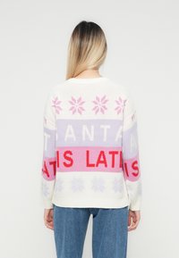 Frau mit glattem blonden Haar, die einen weißen Strickpullover mit pinken und roten Schneeflockenmuster sowie Text trägt, kombiniert mit blauen Jeans und in die entgegengesetzte Richtung zeigt.