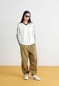 Balta garā piedurkņu blūze ar melnu V veida kaklu un sānu svītrām, apvienota ar brīviem khaki biksēm un baltām sporta kurpēm, uz neitrāla fona.