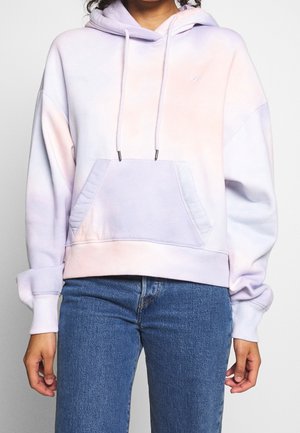 Osoba ubrana w pastelową bluzę z kapturem tie-dye z przednią kieszenią i sznurkami, w połączeniu z niebieskimi dżinsami, stojąca na jednolitym tle.