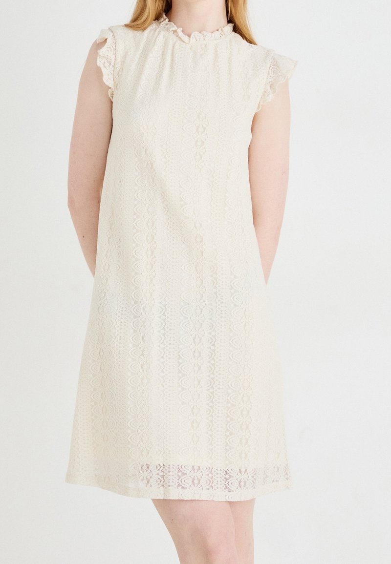 Femme portant une robe en dentelle crème longueur genou avec manches courtes et col volanté sur fond blanc.