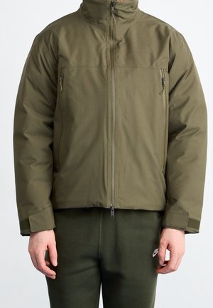 Person trägt eine olivgrüne Jacke mit Reißverschluss, hohem Kragen und Reißverschlusstaschen auf der Brust, kombiniert mit dunkelgrünen Nike-Hosen.