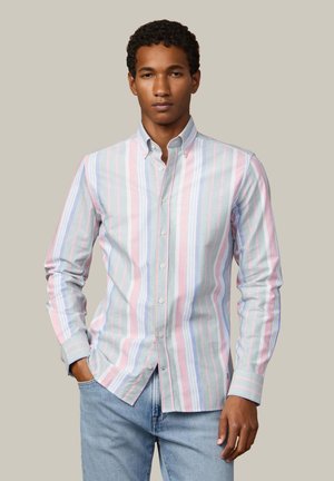 Junger Mann, der ein pastellfarben gestreiftes Hemd mit Knöpfen und hellblaue Jeans trägt, steht mit einer Hand in der Tasche vor einem einfarbigen Hintergrund.