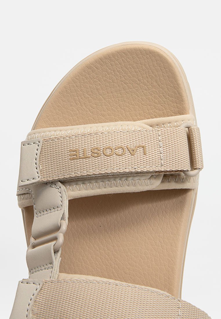 Sandale beige avec un semelle texturée, large sangle avec boucle et le nom de la marque embossé sur la sangle. Matériau doux et flexible.