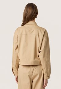 Beige cropped jakke lavet af glat stof med krave og knapmanchetter, med en broderet logo detalje på ryggen.
