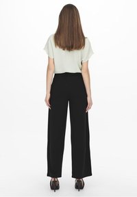 Femme aux cheveux bruns raides se tient de dos, portant un haut léger à manches courtes, un pantalon large noir et des talons hauts noirs.