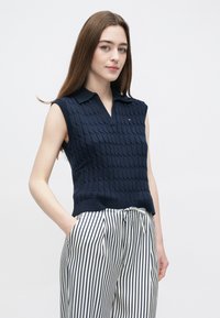 Mornarski pleteni prsluk bez rukava s teksturiranim uzorkom, s ovratnikom u obliku slova V. Uparen s bijelim i mornarskim hlačama s okomitim prugama.