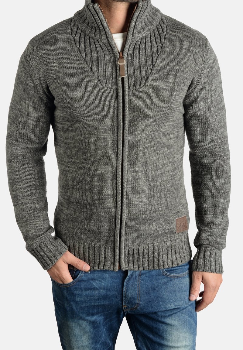 Homme portant un pull gris en tricot zippé avec col, poignets et ourlet côtelés, associé à un jean bleu délavé, mains dans les poches.