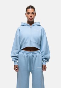 Felpa cropped blu chiaro con zip e cappuccio, abbinata a pantaloni joggers coordinati. Tessuto morbido, vestibilità comoda e dettagli del logo bianco su entrambi i capi.