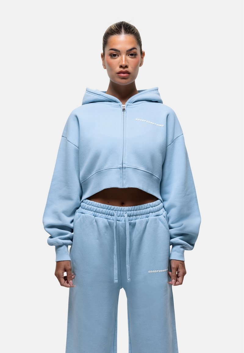 Felpa cropped blu chiaro con zip e cappuccio, abbinata a pantaloni joggers coordinati. Tessuto morbido, vestibilità comoda e dettagli del logo bianco su entrambi i capi.