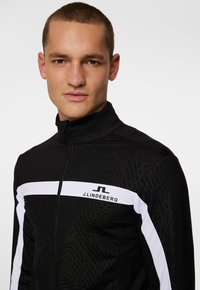 Black zip-up jacket with high collar, featuring a white horizontal stripe and logo, smooth texture with a subtle pattern throughout.