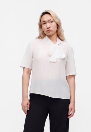 Jong persoon met lichtblond haar, die een witte blouse met korte mouwen, een strikdas en zwarte broek draagt tegen een effen achtergrond.