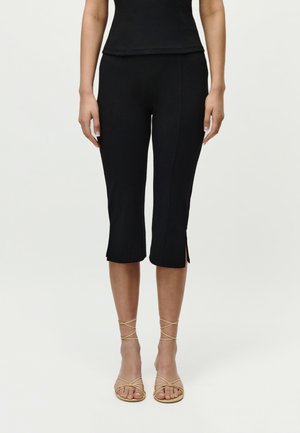 Kvinde iført sorte korte bukser med små slidser ved kanten og beige sandaler med stropper, stående foran en lys baggrund.