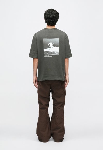 Muškarac stoji okrenut leđima, noseći tamno sivi majicu s grafikom skijaša i smeđe široke hlače, na jednostavnoj pozadini.