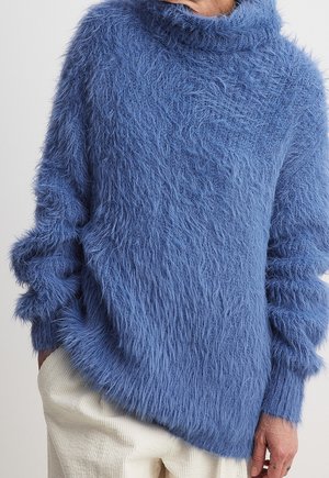 Person iført en langærmet, blød blå sweater med høj hals og hvide bukser, hånd hvilende i lommen.