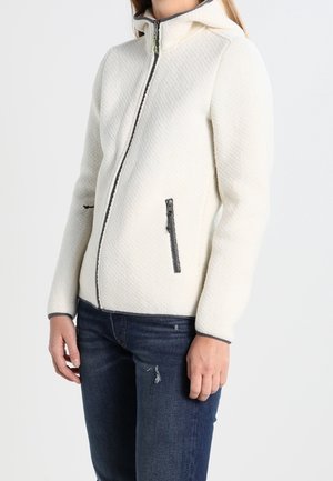 Kvinde iført en hvid quiltet lynlåstrøje med hætte og mørke lynlåsdækning, kombineret med mørkeblå jeans.