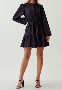 Abito nero con maniche lunghe, chiusura frontale con bottoni, vita elastica e orlo a volant a strati. Tessuto liscio con silhouette strutturata.