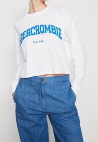 Weißes, langärmeliges cropped Shirt mit blauer Schrift "Abercrombie New York", kombiniert mit blauen, hochgeschnittenen Denim-Hosen.