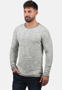 Pull gris à manches longues avec un motif de tricot texturé, encolure ronde et poignets contrastés. Étiquette de marque marron en bas.