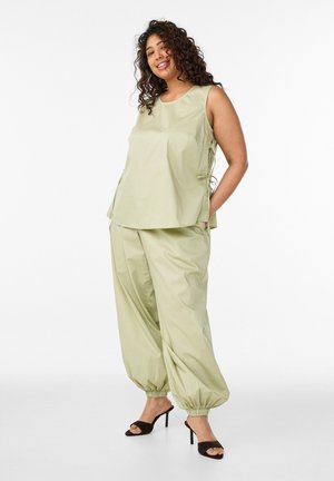 Žena s kučeravými vlasmi oblečená v svetlozelenom tielku bez rukávov a voľných nohaviciach s elastickými manžetami a čiernych sandáloch na podpätku, stojaca.