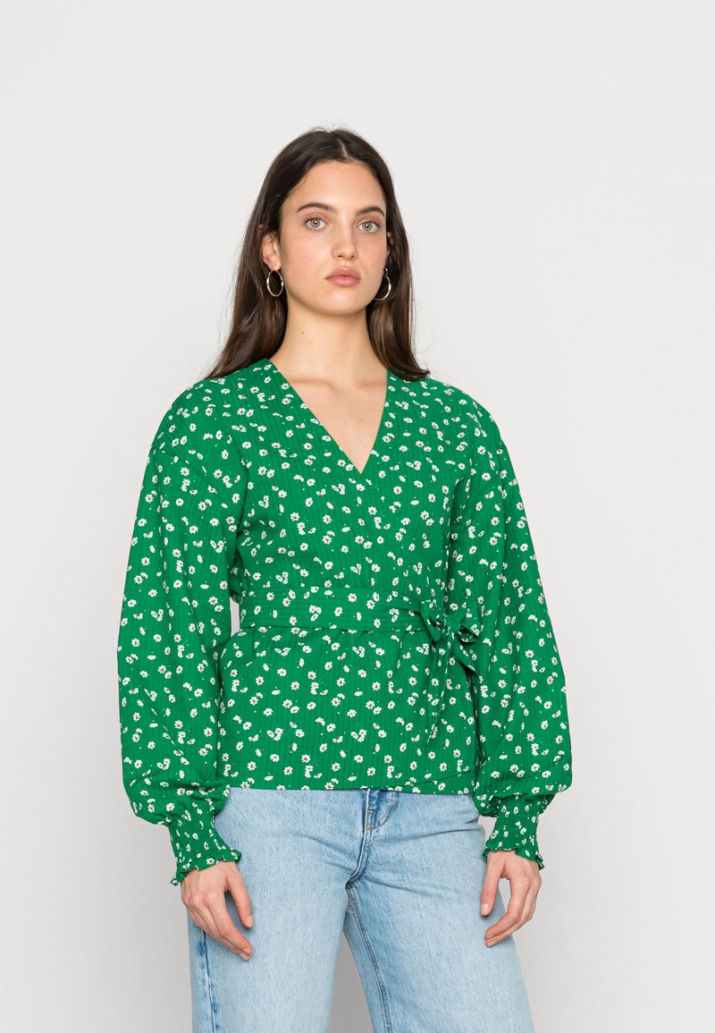 Blusa floral verde com decote em V, mangas longas bufantes e cintura atada. Apresenta padrões florais brancos e tecido texturado. Combinada com jeans claros.