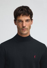 Svart polotröja i mjukt tyg, med en slimmad passform och ribbad halsringning, samt en liten röd logotypdetalj på bröstet.