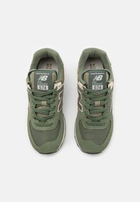 Zielone sneakersy New Balance 574 mają siateczkowy i zamszowy design, sznurowane zapięcie, wyściełaną podeszwę oraz wyraźne beżowe logo „N”.
