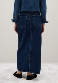 Gonna lunga in denim con lavaggio blu scuro, caratterizzata da due tasche posteriori, una cucitura centrale e una silhouette dritta. Modello che indossa delle scarpe basse nere.