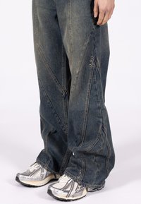 Luźne niebieskie dżinsy z ukośnymi szwami i metalowymi oczkami na mankietach, w połączeniu ze srebrno-białymi butami sportowymi.