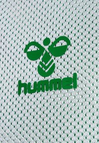 Logo verde su tessuto a rete bianco con un motivo a diamante ripetuto, con il nome del marchio "hummel" ben visibile.