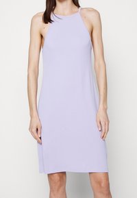 Vrouw in een mouwloze, knie-lange lila jurk met dunne schouderbandjes en een hoge halslijn tegen een effen witte achtergrond.