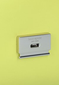 Ferramenta rettangolare in argento con inciso il testo "kate spade NEW YORK", montata su uno sfondo giallo brillante e testurizzato.