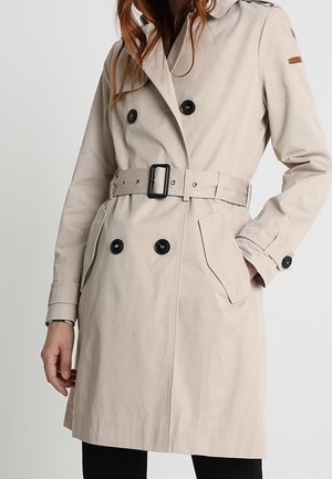 Beżowy dwurzędowy płaszcz trencz z czarnymi guzikami, paskiem w talii, kieszeniami z patkami oraz paskami przy mankietach noszony przez żeńską modelkę.