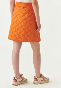 Falda acolchada naranja con un diseño entallado y acabado texturizado. Presenta cremallera lateral y cae por encima de la rodilla. Llevada con zapatillas beige y rojas.