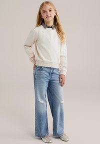 Felpa rosa chiaro con scollatura a V, indossata sopra una camicia bianca e nera con colletto, abbinata a jeans larghi e strappati di colore blu.