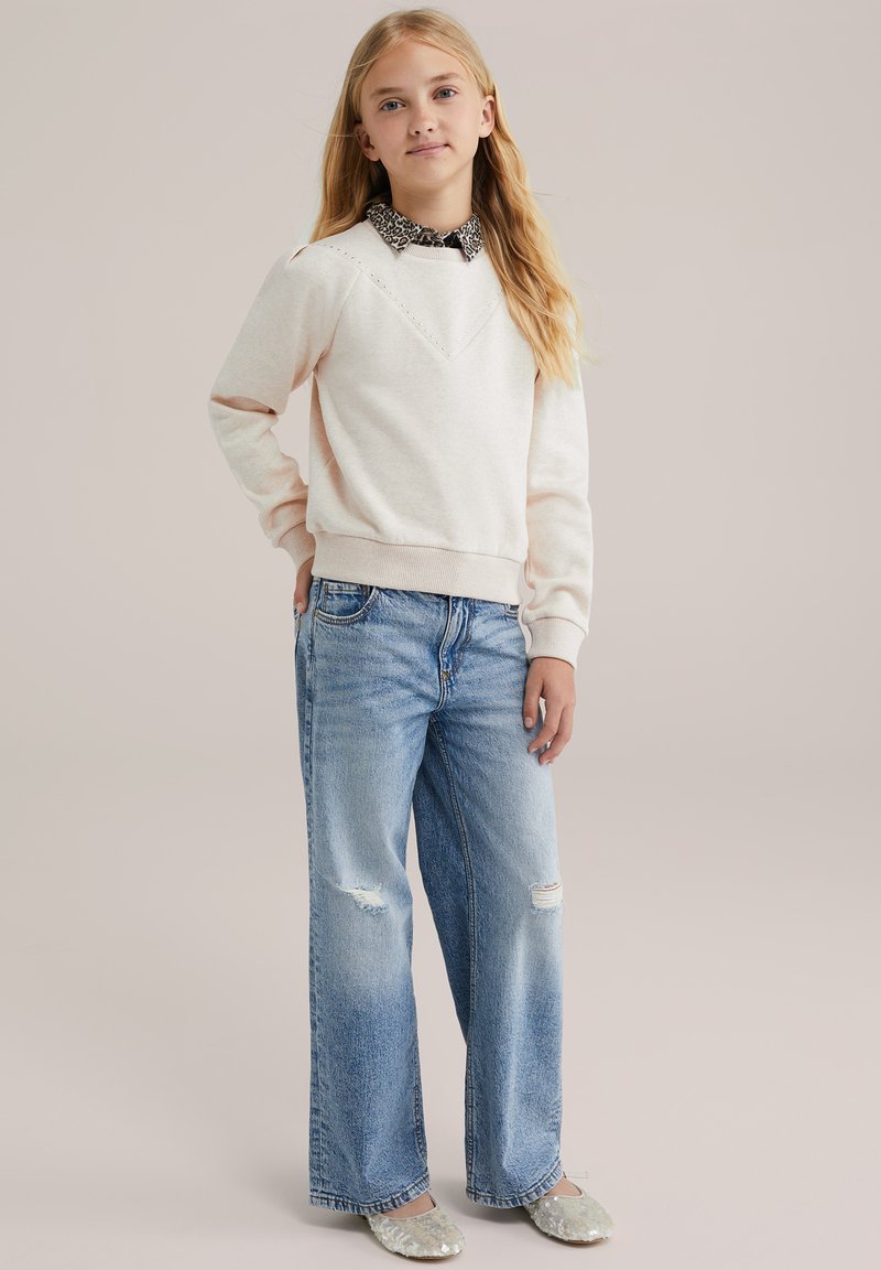 Felpa rosa chiaro con scollatura a V, indossata sopra una camicia bianca e nera con colletto, abbinata a jeans larghi e strappati di colore blu.