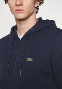 Hanorac cu fermoar de culoare bleumarin, fabricat dintr-un material moale, având o glugă cu șnur și logo-ul brodat al unui crocodil verde pe piept.