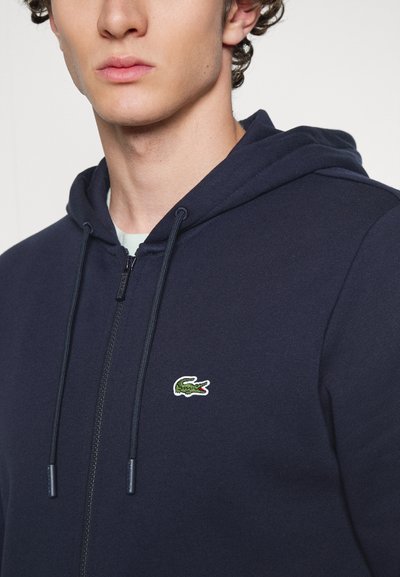 Hoodie zippé marine en tissu doux, doté d'une capuche avec cordon de serrage et d'un logo de crocodile vert brodé sur la poitrine.