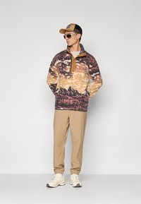 Man met een gemêleerde fleecepullover met bergontwerp, beige broek, off-white sneakers, een tan en zwarte pet, en witte zonnebril, die tegen een witte achtergrond staat.