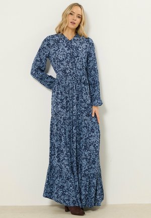 Frau in einem langärmligen, marineblauen Maxikleid mit Blumenmuster, die mit einer Hand in der Hüfte vor einer einfarbigen Wand steht.