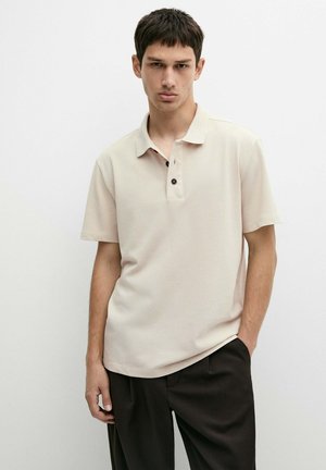 Ung mand med kort, mørkt hår iført beige poloshirt og sorte bukser står med den ene hånd i lommen mod en ensfarvet baggrund.