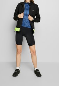 Czarna lekka kurtka z neonowymi zielonymi akcentami, niebieska rozpinana bluzka pod spodem oraz czarne spodenki kolarskie. Obuwie to czarne buty kolarskie.