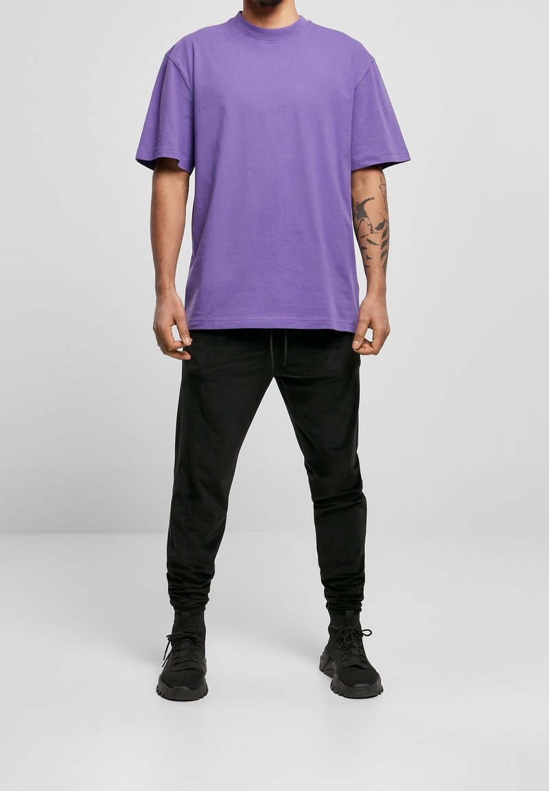 Homme portant un t-shirt violet ample, un pantalon de jogging noir, des baskets noires, et un tatouage sur son avant-bras gauche, debout devant un fond gris.