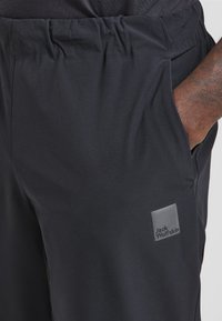 Schwarze Sportshorts mit einem elastischen Bund, ausgestattet mit einer Seitentasche und einem kleinen grauen Logo-Patch mit der Aufschrift "Jack Wolfskin". Glatte Textur.