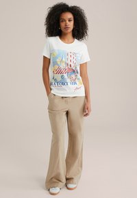 Witte katoenen t-shirt met kleurrijke "La Dolce Vita" grafiek, gecombineerd met beige wijde broek en witte sneakers met lichtbruine zolen.