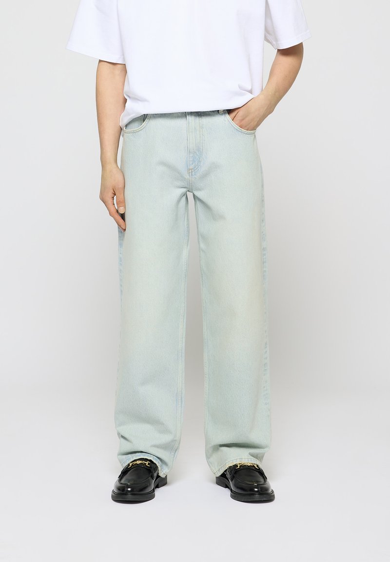 Jeans azzurri chiari a gamba larga con una finitura sbiadita, caratterizzati da un classico modello a cinque tasche. Indossati con una t-shirt bianca semplice e scarpe nere.